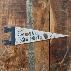 A white triangular flag with blue and orange text and illustrations, including a sewing needle and thread, is displayed on a wooden surface.