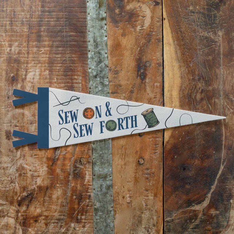 A white triangular flag with blue and orange text and illustrations, including a sewing needle and thread, is displayed on a wooden surface.