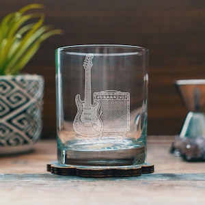 May include: A clear glass tumbler with an etched design of an electric guitar and amplifier. The glass sits on a wooden coaster with a floral design. The background includes a potted plant and a wooden surface.