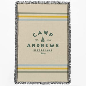Puede incluir: Una manta tejida con fondo crema y rayas verdes, amarillas y azules. En el centro de la manta hay un logotipo que dice "Camp Andrews, Est. 1990, Sebago Lake, Maine".
