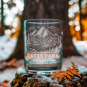 May include: A clear glass tumbler featuring an etched design of a mountain range, a forest, and a cabin near a lake. The text ESTES PARK is engraved in a bold, sans-serif font below the landscape illustration on the front of the glass.