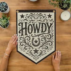 a banner with the word "HOWDY" written on it, surrounded by decorative elements such as stars and a horseshoe. The banner is placed on a wooden surface, with a pair of hands resting on it. There are also some potted plants and a candle in the background.