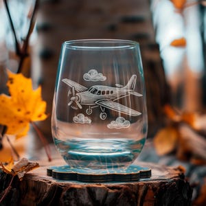 May include: A clear stemless wine glass with an etched design of a propeller airplane and clouds. The glass sits on a blue and brown coaster, placed on a tree stump. The background features fall foliage.