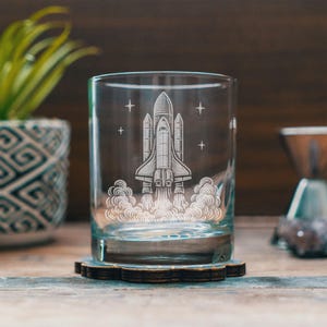 May include: A clear glass tumbler with a detailed etched design of a space shuttle launching, surrounded by stars and smoke. The glass sits on a wooden coaster. The theme is space and exploration.