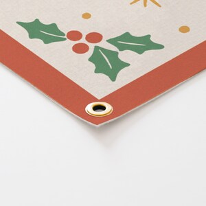 May include: Corner of a banner with a cream background, featuring a red border and gold grommet. The design includes green holly leaves, red berries, and gold starbursts and dots, evoking a festive holiday theme.