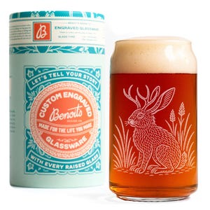 May include: A custom engraved beer glass with a hare design, filled with beer and foam. The glass is next to a light blue cylinder with text that reads "Let's tell your story" and "Custom Engraved Glassware".