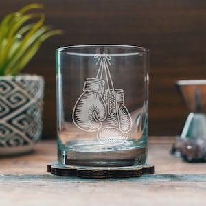 Könnte beinhalten: Ein klares Glas mit einem geätzten Design von Boxhandschuhen, die an einer Schnur hängen. Das Glas steht auf einem dunklen Holzuntersetzer. Im Hintergrund befinden sich eine Topfpflanze und eine Kaffeemaschine.