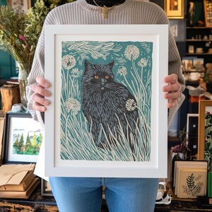 May include: A framed art print featuring a black cat with orange eyes, set against a teal background with white dandelions and tall grass. The print is in a white frame, creating a striking contrast.