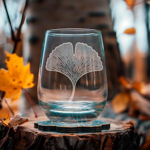 May include: A clear stemless wine glass with a white ginkgo leaf design. The glass sits on a decorative coaster with a blue and gold pattern. The background features fall foliage and a tree stump.