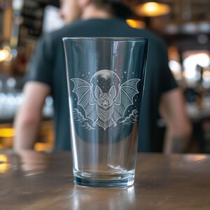 May include: A clear pint glass featuring an etched design of a bat with outstretched wings, a full moon, and clouds. The bat design is centered on the glass. The glass is on a brown surface.