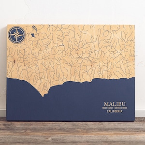 May include: A wooden map of Malibu, California, with blue water and a compass rose in the top left corner. The text "MALIBU" is printed in white at the bottom of the map, along with "WEST COAST - UNITED STATES" and "CALIFORNIA".