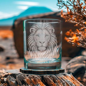 Könnte beinhalten: Ein klares Glas mit einem geätzten Löwenkopf-Design. Die detaillierte Gravur zeigt die Mähne, das Gesicht und die Augen des Löwen. Das Glas steht auf einem Holzuntersetzer. Der Hintergrund umfasst einen verschwommenen Berg und Blätter.