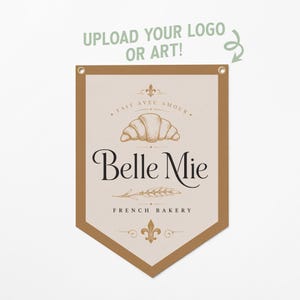 May include: A tan pennant banner featuring a central graphic of a croissant, a wheat stalk, and fleur-de-lis symbols. The text reads "FAIT AVEC AMOUR", "Belle Mie", and "FRENCH BAKERY". Above the banner, the text "UPLOAD YOUR LOGO OR ART!" appears.