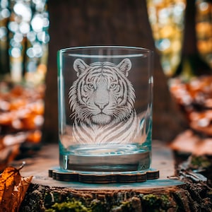 Peut inclure: Un verre transparent avec un motif de tigre gravé. Le visage du tigre est au centre du verre, avec des détails de fourrure et de rayures. Le verre repose sur un dessous de verre en bois, avec un arrière-plan flou de forêt.