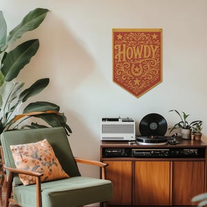 a cozy living room with a green armchair, a wooden cabinet with a turntable and speakers, and a large potted plant. A decorative banner with the word "HOWDY" in an ornate font hangs on the wall above the cabinet.