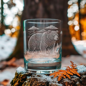 Könnte beinhalten: Ein klares Glas mit einem geätzten Design eines Bären, Bergen und Wolken. Der Bär ist das zentrale Motiv, mit einer Waldszene im Hintergrund. Das Glas steht auf einer moosbewachsenen, schneebedeckten Oberfläche.