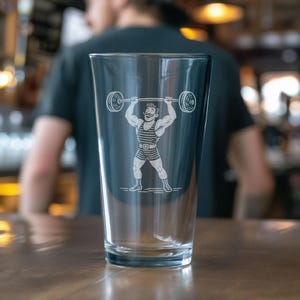 May include: Clear pint glass featuring a vintage illustration of a weightlifter. The design is etched in white and shows a muscular man lifting a barbell overhead. The glass is tapered and sits on a wooden surface, with a blurred background.
