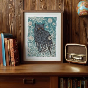May include: A framed art print featuring a black cat with orange eyes, set against a teal background with white dandelions and tall grass. The print is in a brown wooden frame, displayed on a wooden surface.