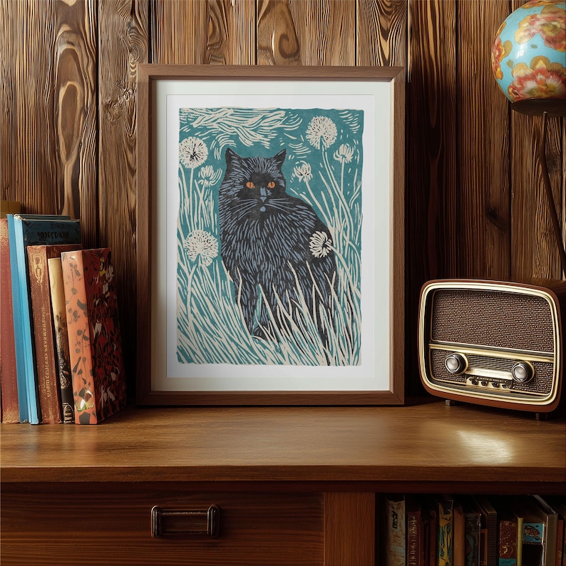 May include: A framed art print featuring a black cat with orange eyes, set against a teal background with white dandelions and tall grass. The print is in a brown wooden frame, displayed on a wooden surface.