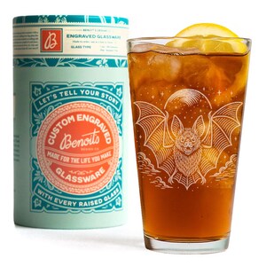 May include: A glass of iced tea with a lemon wedge, decorated with a bat illustration. Beside it is a cylindrical container with the text "Custom Engraved Glassware" and "Let's Tell Your Story."