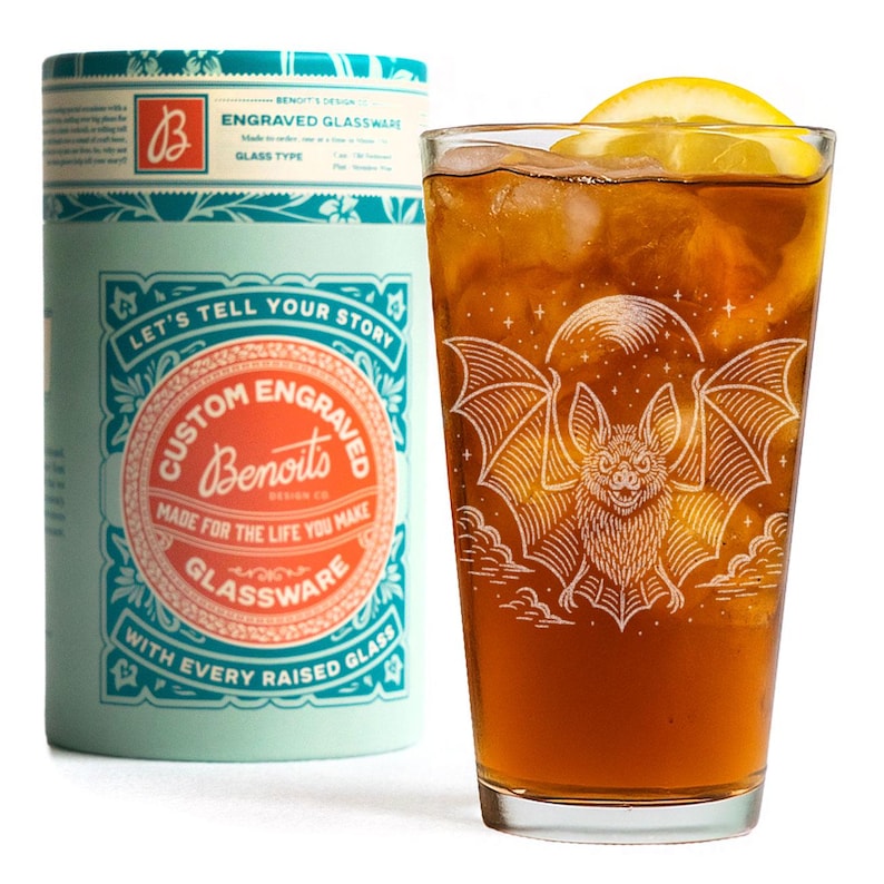 May include: A glass of iced tea with a lemon wedge, decorated with a bat illustration. Beside it is a cylindrical container with the text "Custom Engraved Glassware" and "Let's Tell Your Story."