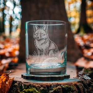 May include: A clear glass tumbler with a detailed etched image of a fox sitting in grass. The fox design is in white, and the glass sits on a wooden coaster. The background features a blurred forest scene with fall foliage.