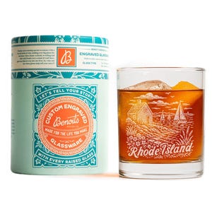 May include: A clear glass tumbler filled with an amber liquid and ice, featuring a white engraved illustration of a coastal scene with a house, sailboat, and the text Rhode Island. Beside it sits a cylindrical light blue and orange cardboard tube.