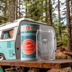 May include: A clear glass can-shaped drinking glass with a bat and moon design, beside a cylindrical container with the text "Custom Engraved Glassware". The items are on a wooden table with a vintage camper in the background.