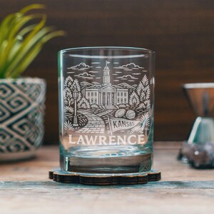 Könnte beinhalten: Ein klares Glas mit einem detaillierten geätzten Design, das ein Gebäude, Bäume und das Wort "Lawrence" darunter zeigt. Das Wort "Kansas" ist ebenfalls auf dem Glas geätzt. Das Glas steht auf einem Holzuntersetzer.