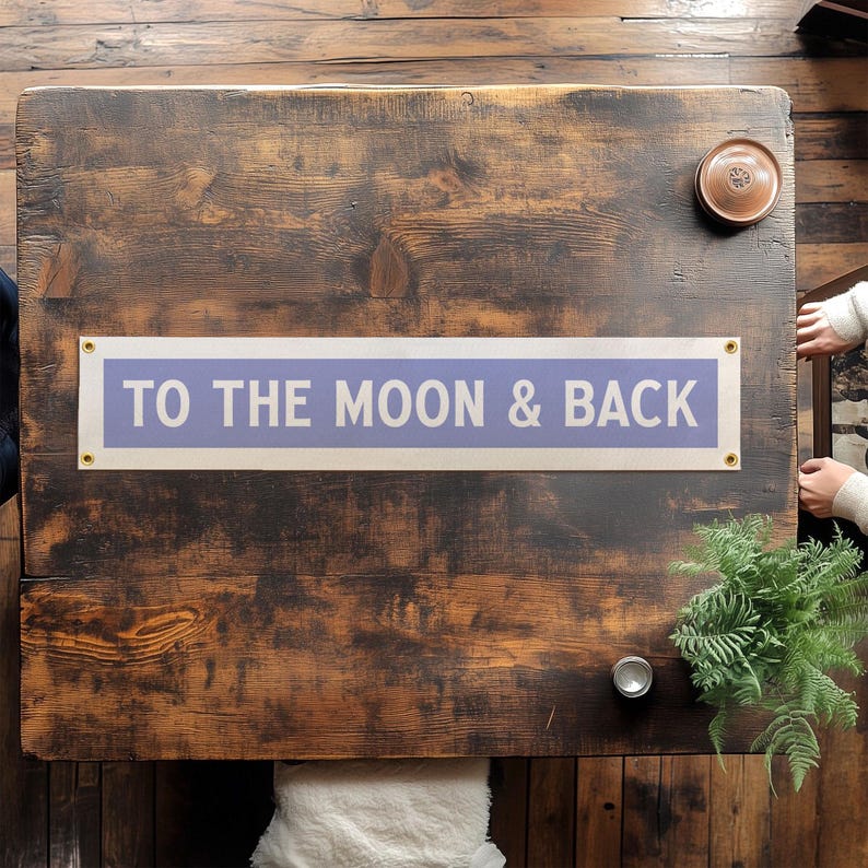May include: A rectangular sign with the text "TO THE MOON & BACK" in white letters on a light purple background. The sign is mounted on a dark brown wooden surface. The sign has gold-colored grommets in each corner.