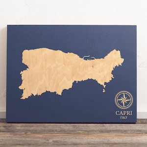Op de afbeelding: Een houten kaart van het eiland Capri, Italië, op een donkerblauwe achtergrond. De kaart is uit hout gesneden en heeft een kompasroos in de rechterbenedenhoek. De tekst "CAPRI ITALIË" staat onder de kompasroos.