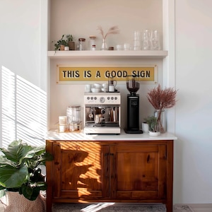 Può includere: Una postazione per il caffè domestica con una macchina per espresso bianca, un macinacaffè nero e un cartello con la scritta "THIS IS A GOOD SIGN". La postazione è posizionata su un mobile in legno con un piano di lavoro bianco ed è decorata con piante e vetreria.