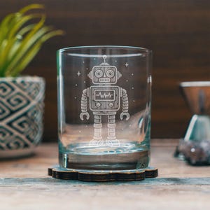 May include: A clear glass tumbler with a detailed etched design of a robot. The robot has circular eyes, a rectangular chest with a heartbeat line, and segmented arms and legs. The glass sits on a dark wooden coaster.