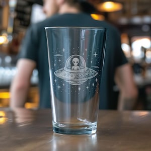 May include: A clear pint glass with an etched design of a UFO and an alien. The alien is in the center of the UFO, with stars surrounding the ship. The glass is on a wooden surface.