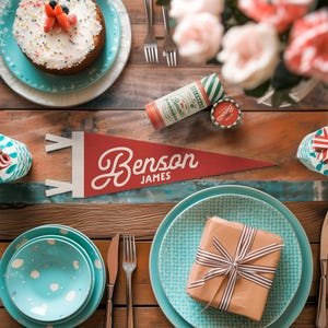 Puede incluir: Una mesa festiva con un pastel, platos y un regalo. Se muestra un banderín rojo con "Benson James" en escritura blanca. Otros elementos incluyen un recuerdo de fiesta, cubiertos y rosas rosas.
