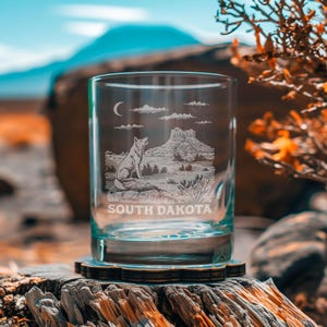 May include: A clear glass tumbler with an etched design featuring a wolf, a mountain landscape, clouds, and a crescent moon. The words "SOUTH DAKOTA" are etched below the image. The glass sits on a wooden coaster.