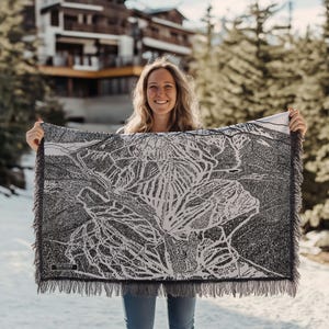 May include: A woman is holding up a white and black woven throw blanket with a detailed map of a ski resort printed on it. The map shows the ski runs, lifts, and other features of the resort. The text "Sun Peaks" is printed in the bottom left corner of the blanket.