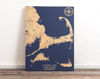 Op de afbeelding: Een donkerblauwe houten kaart van Cape Cod, Massachusetts, met een gouden kompasroos in de rechterbovenhoek. De kaart is gedetailleerd met gouden lijnen die de waterwegen en de kustlijn voorstellen.