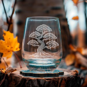May include: A clear stemless wine glass with an etched design of a bonsai tree. The glass sits on a decorative coaster with a blue and gold pattern, placed on a tree stump. The background features fall foliage.