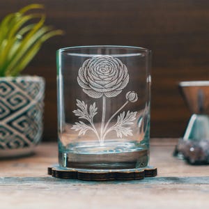May include: A clear glass tumbler with a detailed etched floral design of a ranunculus flower. The glass sits on a dark wooden coaster with a scalloped edge. The background is blurred, suggesting a rustic setting.