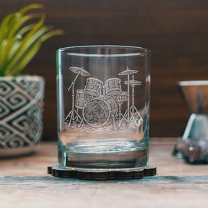 May include: A clear, cylindrical glass with an etched design of a drum set. The drum set includes a bass drum, snare drums, and cymbals. The glass sits on a wooden coaster with a floral design. The background is a blurred wooden surface.
