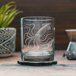 May include: A clear glass tumbler with an etched design of a squid. The squid illustration is white and detailed, featuring tentacles and body markings. The glass sits on a dark wooden coaster. The background includes a potted plant and a coffee maker.