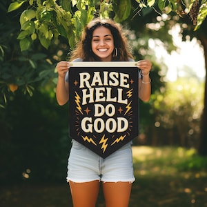 May include: A black pennant with the words "RAISE HELL DO GOOD" in cream-colored, bold lettering. The pennant has gold lightning bolt and star accents. The pennant is held up by a person in a natural outdoor setting.