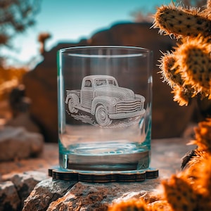 May include: A clear glass tumbler featuring an etched design of a classic pickup truck. The glass rests on a decorative coaster, set against a desert backdrop. The truck design is detailed and prominent.