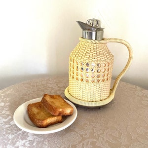 May include: A wicker-wrapped thermos with a silver lid and handle, sitting on a table with two slices of toasted bread on a white plate.