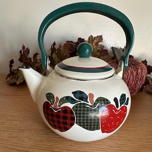May include: White enamel teapot with a green handle and lid knob. The teapot features apple designs in red plaid, green houndstooth, and red with white dots. A red and green stripe decorates the lid. The teapot is a kitchenware item.