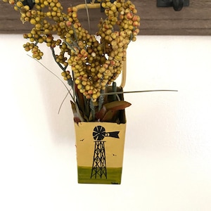 May include: A small yellow metal container with a black windmill design hangs from a hook on a wooden wall. The container is filled with a bouquet of dried yellow berries.