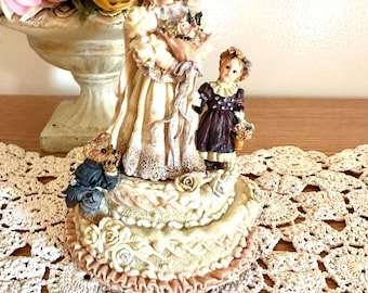 May include: A decorative figurine featuring a larger figure in a cream-coloured dress and hat holding a bouquet, a smaller figure in a dark purple dress, and a small teddy bear. The figures stand on a tiered, circular base decorated with sculpted roses.