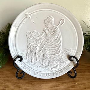 May include: A white ceramic plate with a relief scene of the Nativity, including a star, figures, and sheep. The plate is displayed on a black metal stand. The words "COME LET US ADORE HIM" are inscribed.