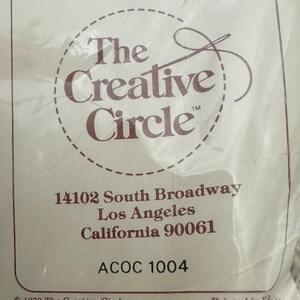 May include: A white label with brown text that reads "The Creative Circle" with a stylized needle and thread logo. The label also includes the address "14102 South Broadway, Los Angeles, California 90061" and the code "ACOC 1004". The bottom of the label reads "&copy; 1979 The Creative Circle" and "Printed in USA."